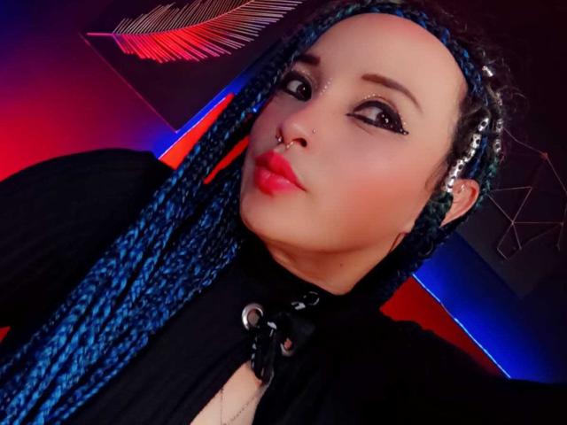 Dalila Musa, con trenzas azules, un aro en la nariz y lápiz labial rojo, vestida con una blusa negra con un aro de metal, mirando a la cámara con una expresión ligeramente fruncida..