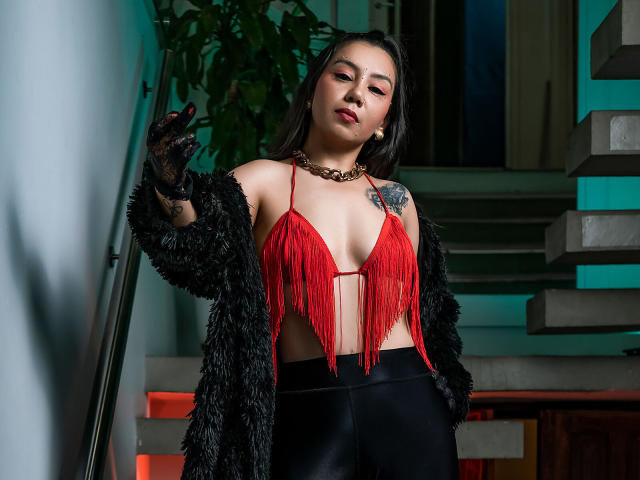 CharlotteJoliie with long dark hair, red fringed top, black fur coat, black pants, black gloves, gold necklace, tattoo on right shoulder, standing on stairs, looking at camera.