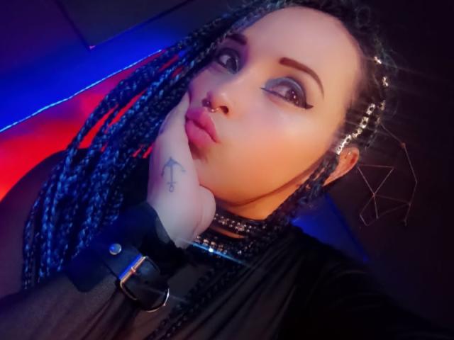 DalilaMusa with braided hair, nose ring, and black choker, pouting with hand on cheek, wearing a black top, blue and red lights in background, tattoo on wrist.