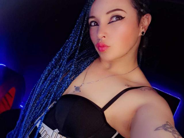 DalilaMusa with long blue braids, black strap dress, pouty pink lips, nose ring, black eyeliner, necklace, tattoo on right arm, looking at camera. Boobs visible in black dress.