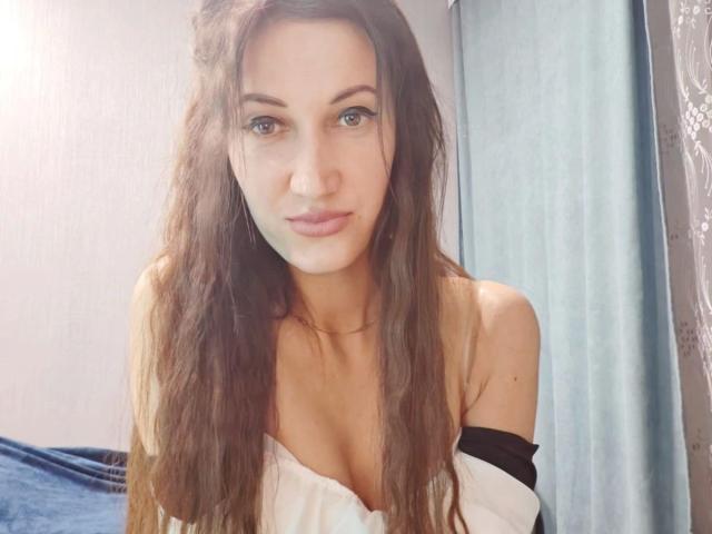 ModestMonika – attraktiv mit langen, braunen, gewellten Haaren, heller Haut, blauen Augen und kleinen Brüsten in einem weißen Top mit einem Schulterteil, einer leicht geöffneten schwarzen Bluse und einem leichten Lächeln..