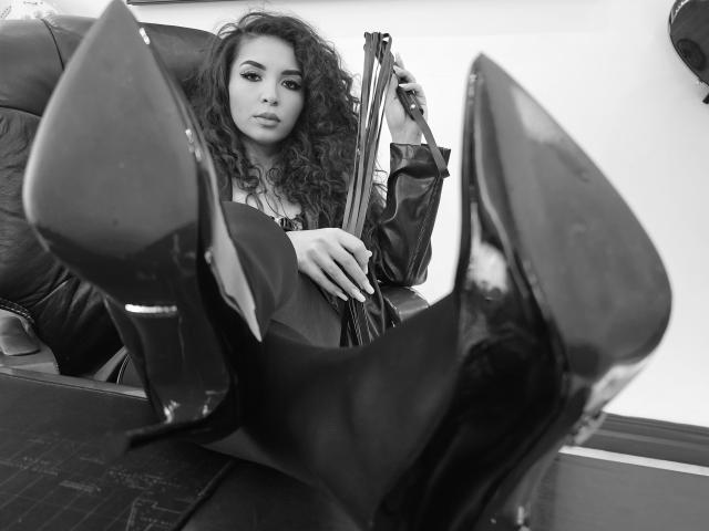 KayaOcean with curly hair, sitting, wearing a leather jacket, black high heels, showing pointed toes, holding a rifle, looking at the camera, black and white image.