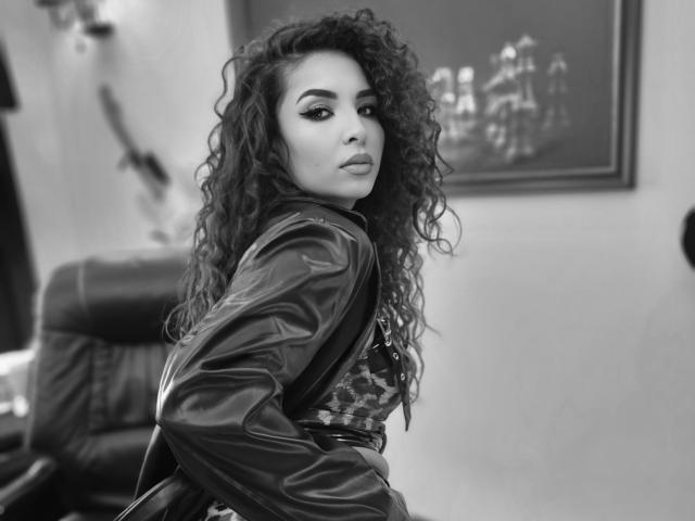 KayaOcean with curly hair, wearing a leather jacket and camo top, looking over her shoulder, black-and-white photo, showing her pouty face and defined eyebrows.