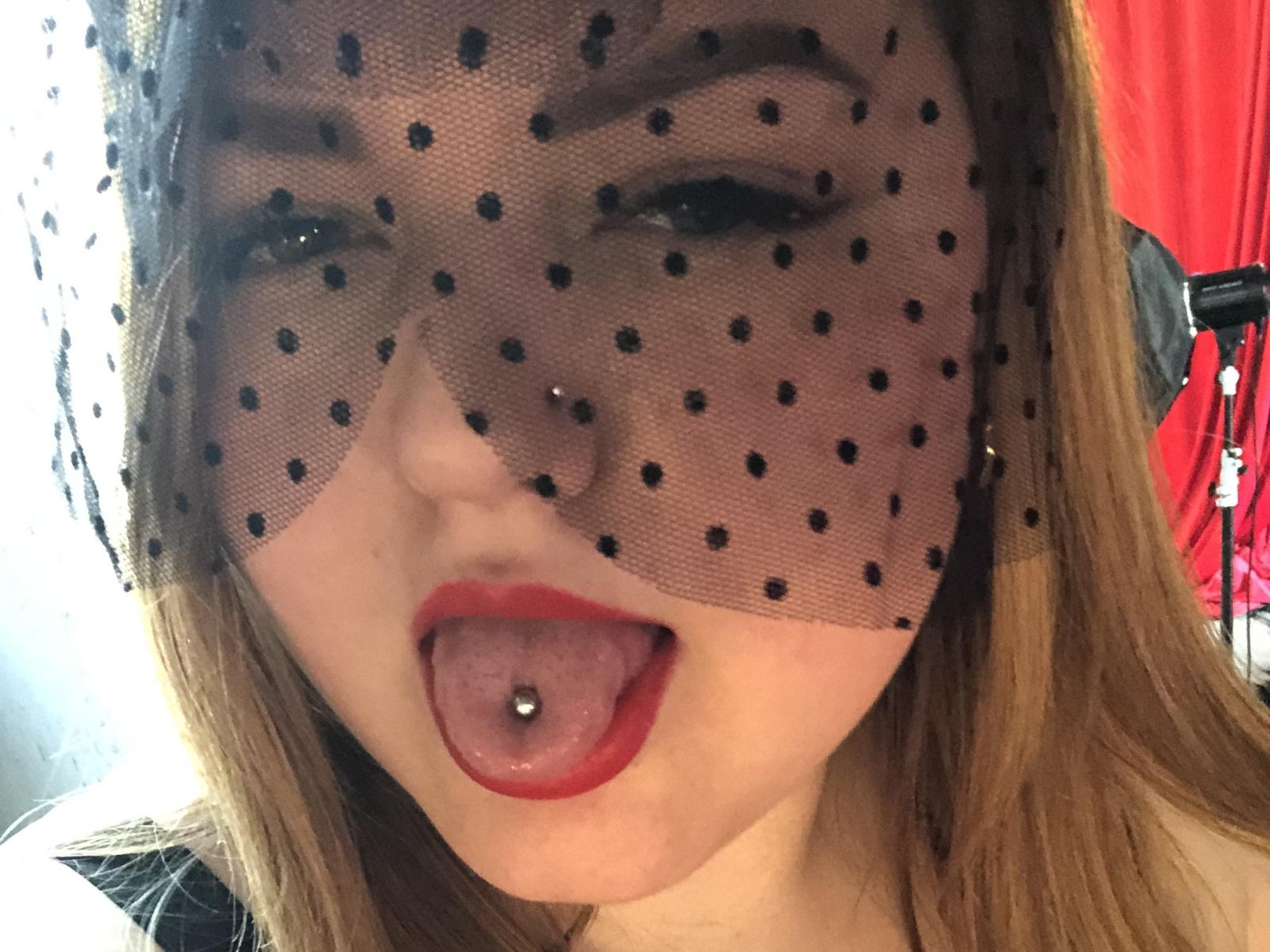 AdelXPeach, avec un masque à pois, la langue tirée, du rouge à lèvres, et un piercing sur la langue. Elle a la peau claire, les cheveux bruns, et un piercing au nez. Elle n'a pas de seins dans cette photo..