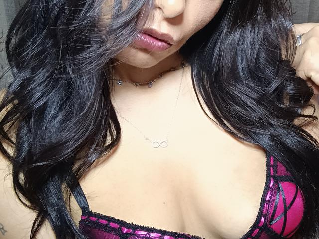 CherryySweet has dark wavy hair, pink and black lace bra, and a silver necklace with a circular pendant. Her pouty, pink-lipned face is partially visible. Boobs are prominent.