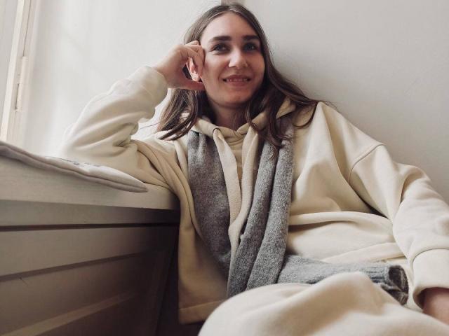 HottieBu with long brown hair, smiling, wearing a beige hoodie and gray scarf, leaning on a windowsill, hand under chin, looking relaxed and cute.