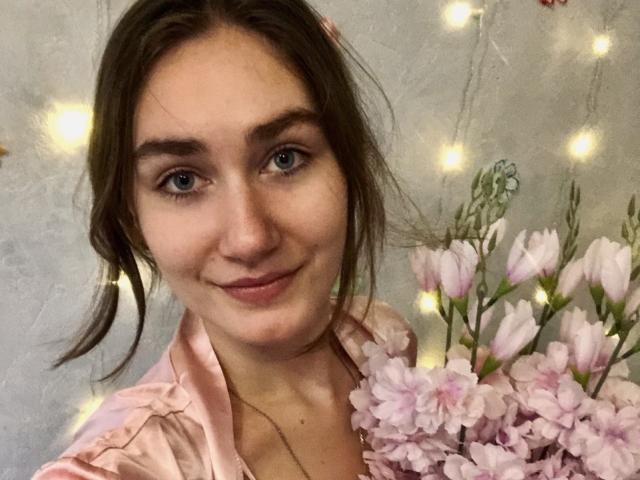 HottieBu with blue eyes, brown hair in pigtails, pink shirt, holding pink flowers, smiling softly. Fairy lights in background. Cute and pretty.