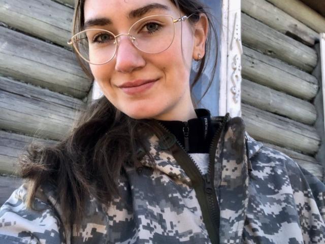 HottieBu with glasses, gray camo jacket, and wavy brown hair, smiling at the camera, showing her cute nose and small earrings.