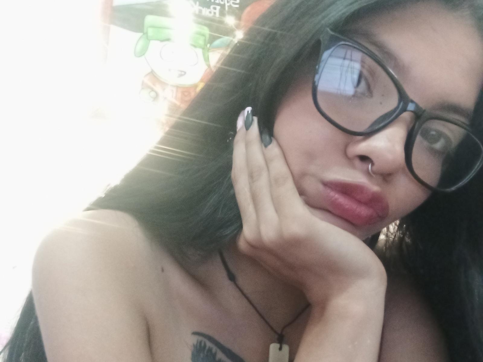 EvilEvaHot with black glasses, nose ring, and red lipstick, pouting with hand on cheek, black nails, and a tattoo above her tits, wearing a necklace.