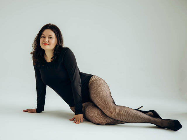 LolaHikls with dark curly hair, black long-sleeve top, fishnet pantyhose, and black heels, lying on white floor, arms and legs spread, smiling with red lips.