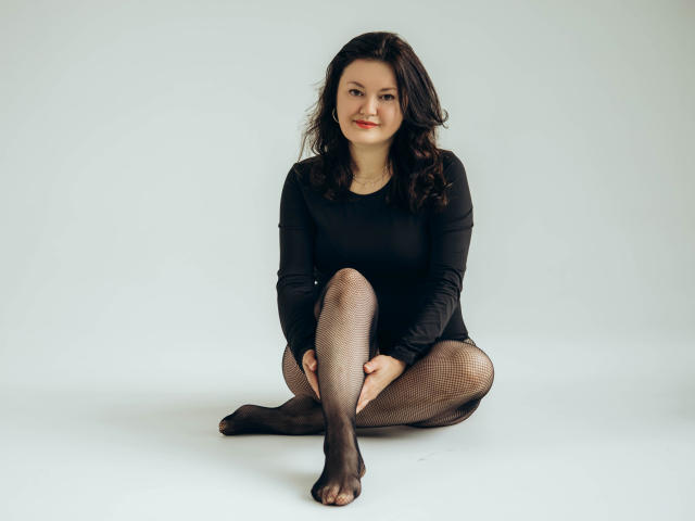 LolaHikls with wavy dark hair, wearing a black long-sleeve top and fishnet stockings, sitting with legs crossed, smiling, and showing a cute face. Boobs, ass, pussy, visible.