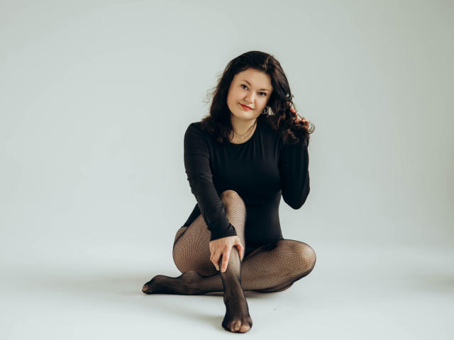 LolaHikls has dark curly hair, wearing a black long-sleeve top and fishnet tights, sitting on the floor with one hand in her hair, smiling at the camera.