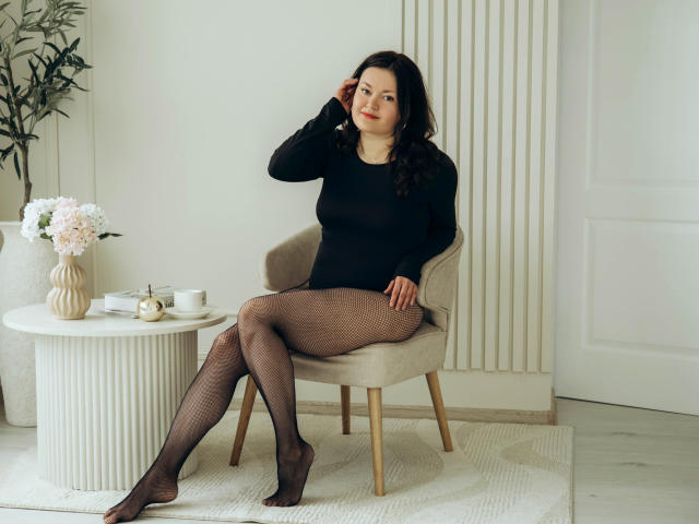 LolaHikls, with dark hair and red lipstick, sits in a beige chair wearing a black sheer top and fishnet leggings, touching her head with a slight smile.