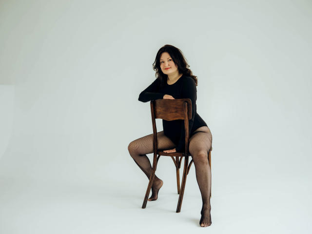 LolaHikls is sitting on a wooden chair, wearing a black long-sleeve top, sheer black pantyhose, and no underwear, with her ass slightly exposed and a relaxed smile.