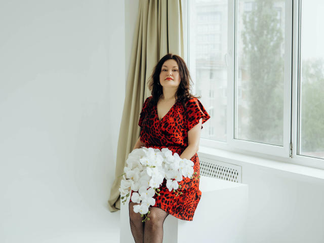 LolaHikls with curly brown hair, red leopard print dress, holding white flowers, sitting by window, beige curtain, white wall, and radiator, looking slightly up, with red lipstick.