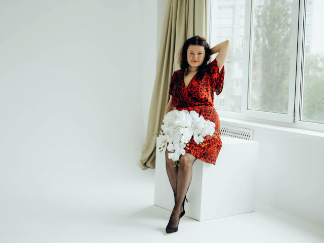 LolaHikls in a red leopard dress, black stockings, and black heels, holding white flowers, sitting on a white cube, near a window, with beige curtains.