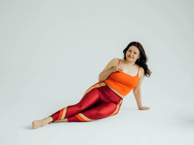 LolaHikls is a curvy woman with large boobs, wearing an orange crop top and red metallic pants, sitting on the floor with a relaxed, smiling expression.
