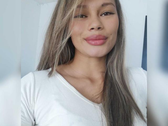 SophiaFoxxy with long, straight brown hair, light brown skin, pink lips, and a small beauty mark on her right cheek, wearing a white ribbed shirt.