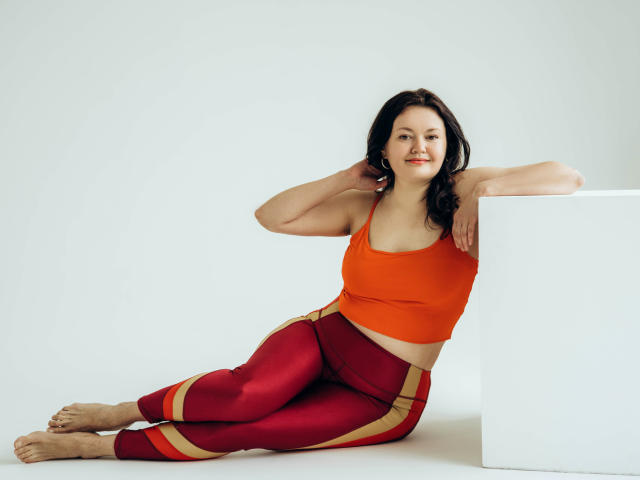 LolaHikls with black hair, orange sports bra, and red pants with yellow stripes, leaning on a white cube, smiling, barefoot, hands in hair, relaxed, attractive.