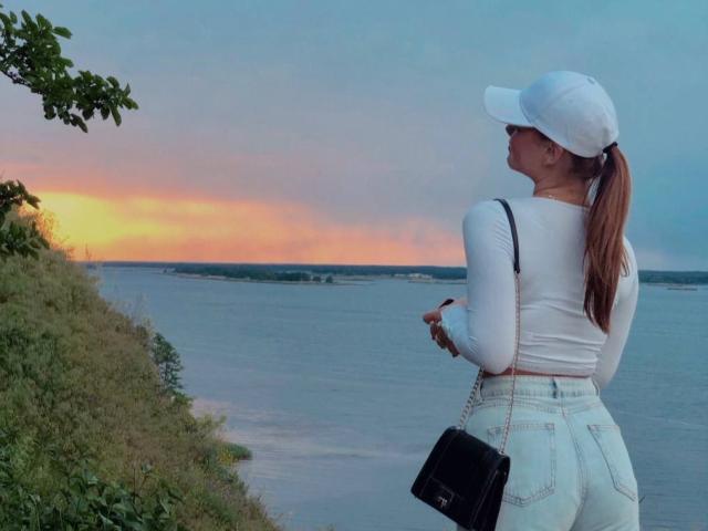 EyeEgida with white cap, white long-sleeve, and high-waisted jeans, holding phone, black purse, standing by river at sunset, green foliage on left, brown ponytail.
