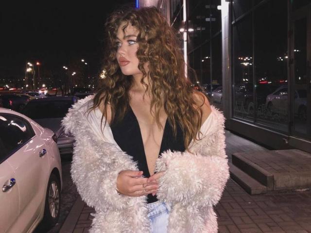 EyeEgida with curly brown hair, wearing a fluffy white jacket, black deep V top, exposing her tits, standing next to a white car at night, looking to the side.