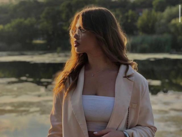 EyeEgida with wavy brown hair, glasses, tan jacket, white tube top, and necklace, standing by a reflective water body, sunlight in her face, looking to the side.