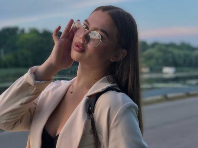 EyeEgida with glasses, pink lips, hand near face, wearing open beige jacket, black top, and necklace, brown hair, outside, dusk, soft light, sexy, pretty.