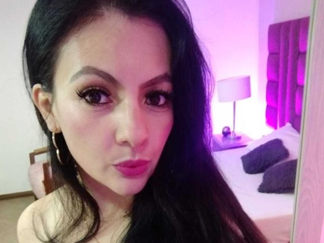 JakyKidman with long black hair, pink lipstick, hoop earrings, and heavy eyeliner, looking at the camera, showing off her tits in a pink-lit bedroom.