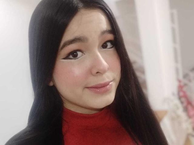 DulceTernura, avec de longs cheveux noirs, portant un pull rouge à col roulé, souriant légèrement avec des lèvres roses brillantes et un maquillage des yeux subtil en forme d'ailes. Elle a un teint clair et un visage mignon..