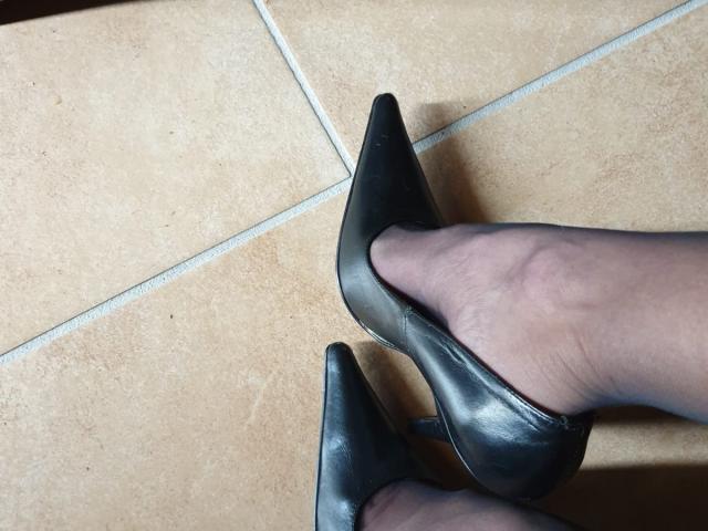 MalenaLoveSex's feet in black, pointy, leather heels with sheer, gray pantyhose, standing on tan, tiled floor. Her toes are slightly curled, showing the heel's texture.