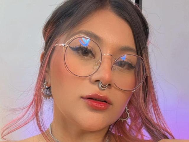 LorraineV with pink-dyed hair, round glasses reflecting a heart, nose ring, and pink lips, wearing earrings, looking straight at the camera. Her skin is smooth and light.