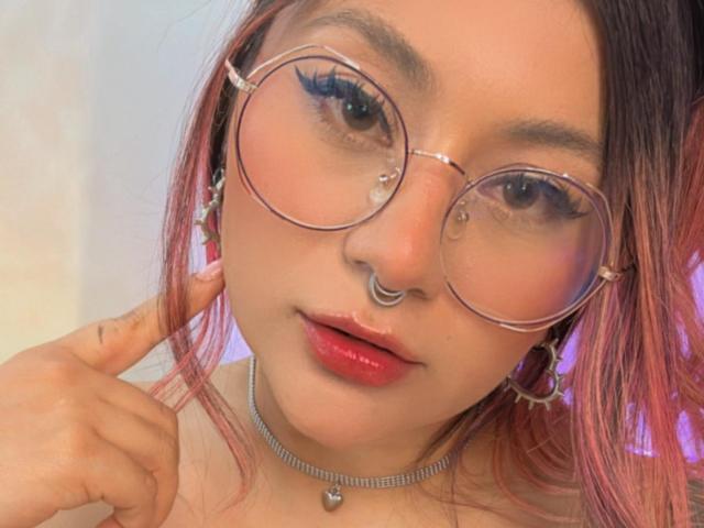 LorraineV with pink-dyed hair, wearing round glasses, nose ring, and red lipstick, touching her ear, has a shiny choker, and looks directly at the camera.