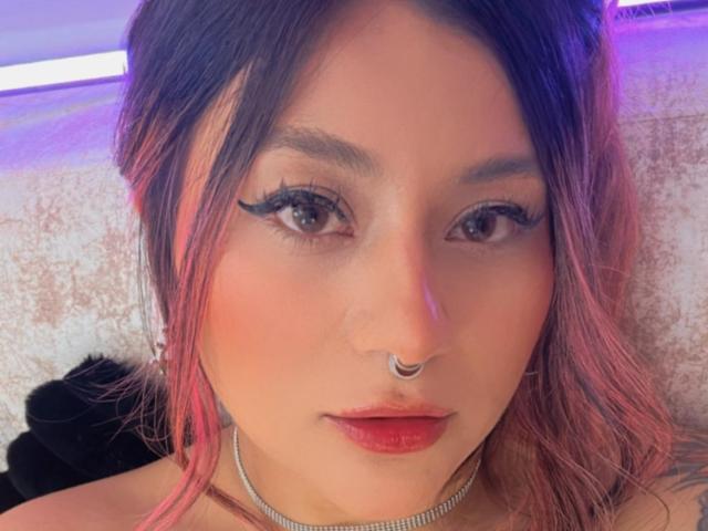 LorraineV has pinkish-brown hair, dark eyebrows, eyeliner, red lipstick, nose ring, choker, and pinkish skin. She's looking at the camera with a slight, sexy, confident smile.