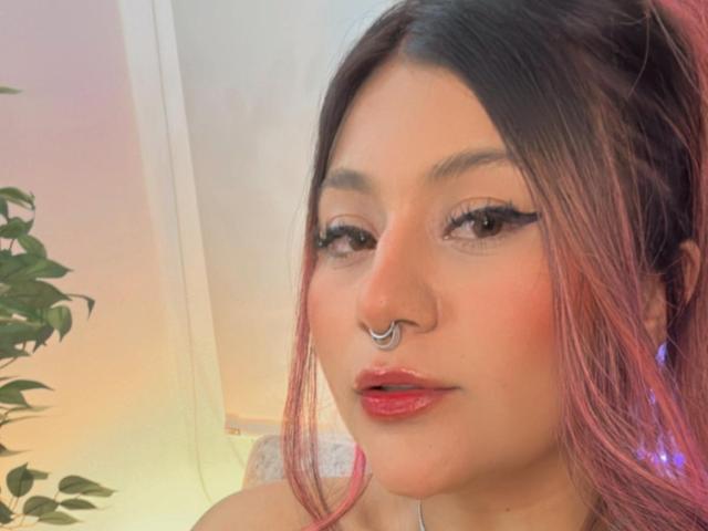 LorraineV with pink-dyed hair, eyeliner, red lipstick, nose ring, and blue earrings, looking at the camera with slightly parted red lips.