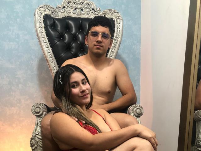 DererAndCleeo with long black hair, red lingerie, and tits sits on a guy's lap. He's shirtless, has curly black hair, and glasses. They're on an ornate black chair.