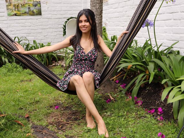 TaylaCooperk, avec de longs cheveux noirs, portant une robe fleurie, assise sur un hamac noir, les jambes croisées, souriante, tenant les cordes du hamac, pieds nus, jardin avec de l'herbe, des fleurs violettes, un mur en briques blanches..
