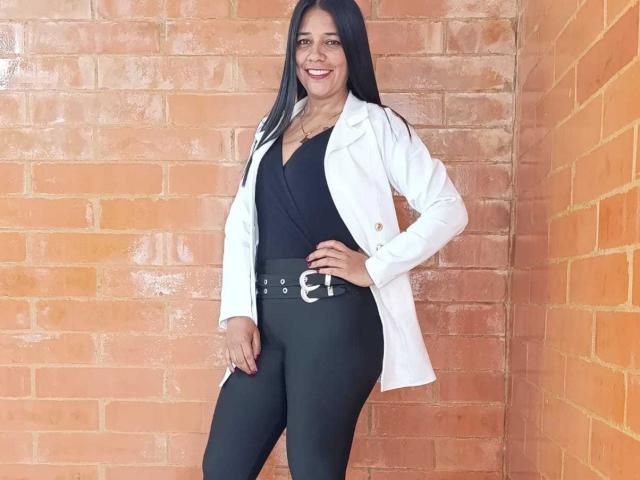 PauletteLoveX, standing, black pants, black top, white coat, belt with buckle, long black hair, smiling, orange brick wall, right hand on hip, left hand down.