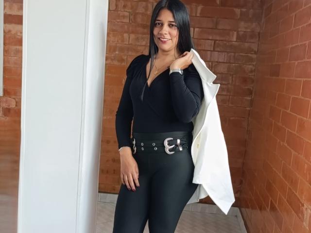 PauletteLoveX with long black hair, smiling, wearing a black V-neck top, black pants with silver belt, holding white jacket, standing in front of red brick wall.