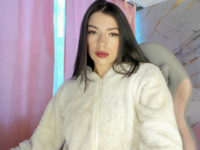 JasmineHypery with long black hair, wearing a fluffy white zip-up, staring at the camera with red lipstick, sitting in a grey chair, looking sexy and confident.