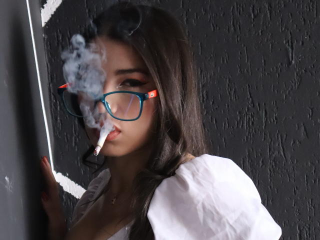 JasmineHypery with long dark hair, blue glasses, and a cigarette, smoking, wearing a white shirt, against a dark textured wall. Her tits are slightly visible.