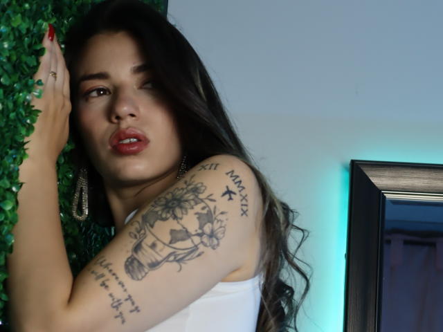 JasmineHypery with long dark hair, white tank top, tattooed arm, red nails, against green wall, looking slightly pouty, standing next to framed mirror with blue light.