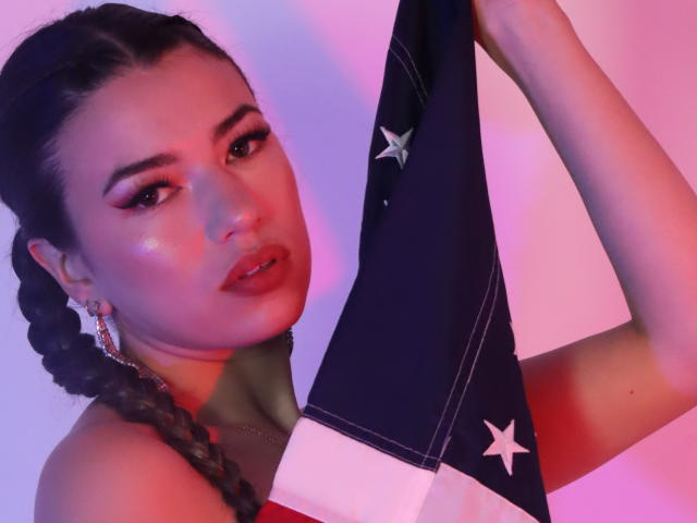 JasmineHypery with braided hair holding a flag, pink and purple lighting, glossy lips, subtle makeup, earrings, raised arms, flag with stars, pink light on right, purple on left.