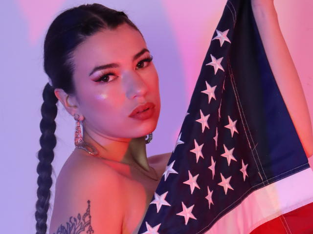 JasmineHypery with dark braided hair, red lipstick, and pink eyeshadow, holding an American flag, showing a tattoo on her shoulder, wearing pink earrings, looking seductively at the camera.