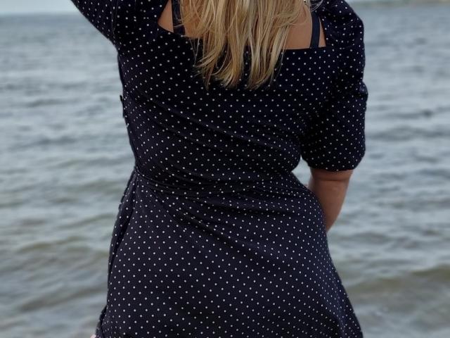 BeatriceSmile's ass in a dark blue polka dot dress with white dots, standing with arms behind her back, facing a rippling water surface, blonde hair visible.