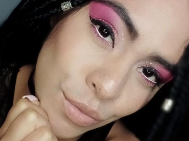 StellaBeckker with pink eyeshadow, black eyeliner, and glitter on her upper eyelids, looking slightly to the side with a subtle smile, holding her chin with her right hand.