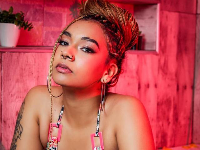MadissonGreey has a nose ring, braided updo, red highlight, and is wearing a colorful halter top, showing a tattoo on her left arm. Red lighting.