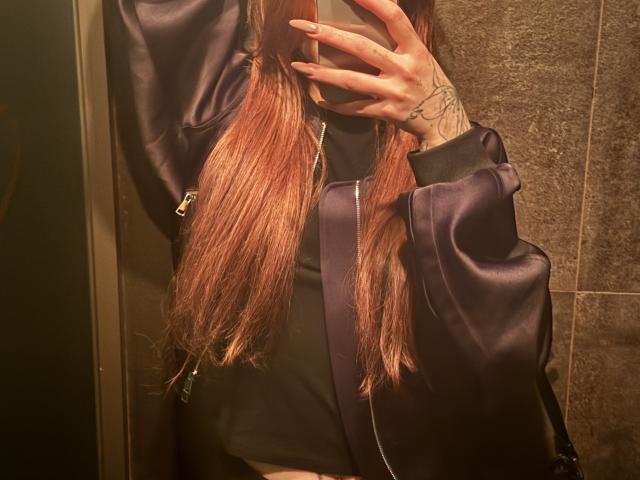 SensualMila with long pinkish-brown hair, wearing a black shirt and purple satin jacket, taking a mirror selfie with her right hand, showing tattoos on her wrist.