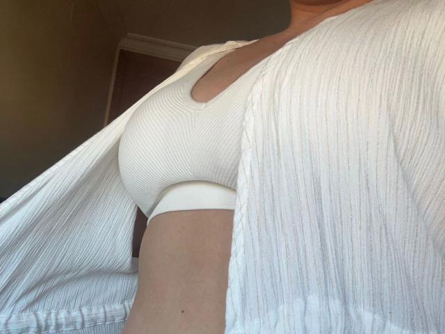 AlexaRoro-hot's tits in a white ribbed shirt, open to reveal a tight white top with a deep neckline, highlighting her chest. Her arm is slightly visible.