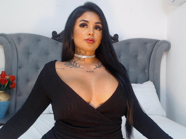 GabrielleKing with long black hair, wearing a black V-neck top, choker, and tattoos on her chest, sitting on a bed with a gray tufted headboard, showing her boobs.