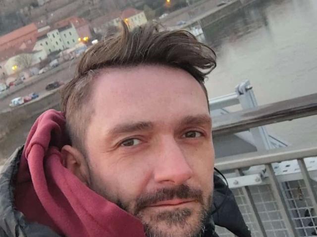 NeueHeimat-hot has a slightly messy brown hairdo, a beard, and is wearing a red hood, looking at the camera with a subtle smile, in a selfie.